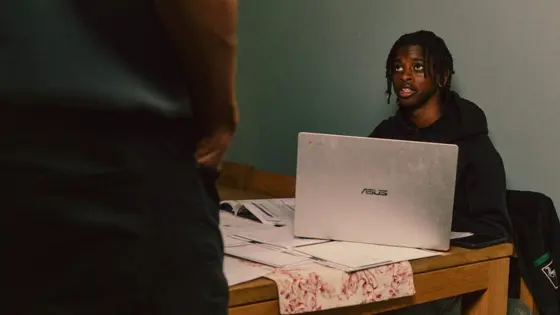 Photograph of a teenager on their laptop, looking up at an adult.