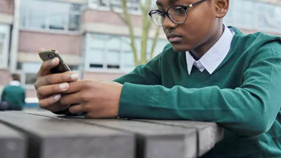 Photo of a teenager sitting outside in their school uniform looking at their phone.
