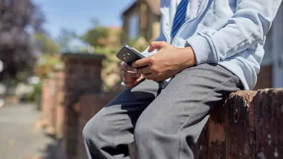 Photo of a teenager sitting outside on a wall while they look at their phone.