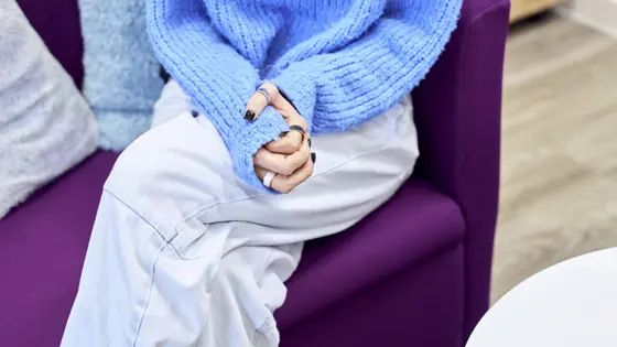 An adult sitting on a sofa at an NSPCC service centre. They are holding their hands on their lap. They look anxious.