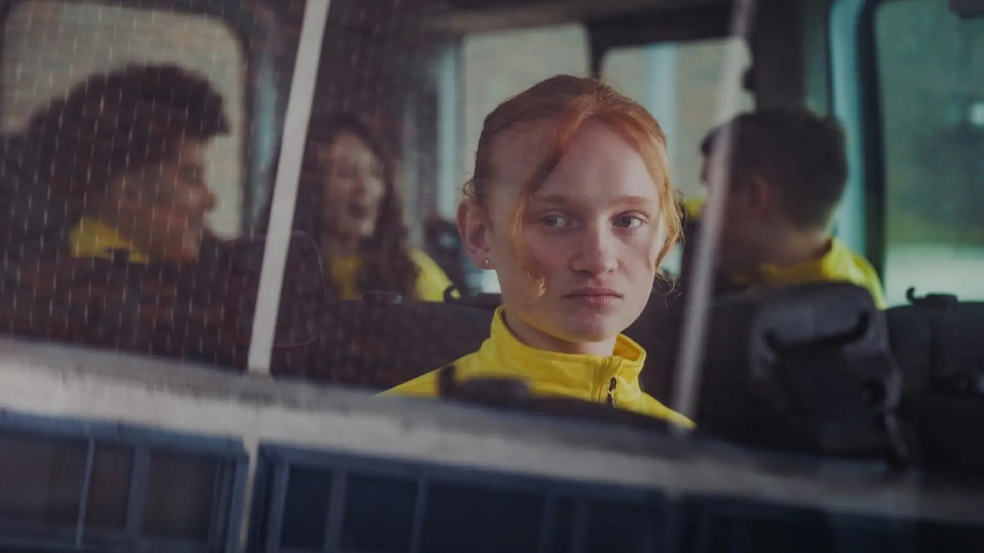 A young girl sat alone on a bus looks out of a window while other children talk in the background.