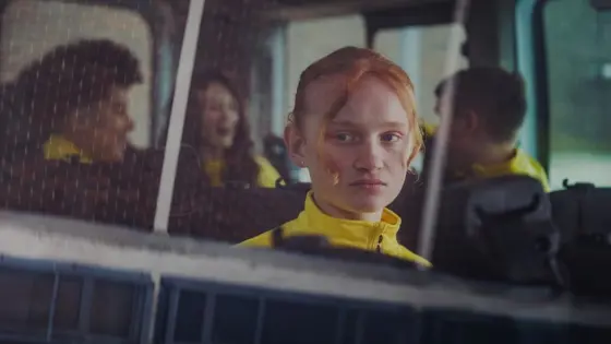 A young girl sat alone on a bus looks out of a window while other children talk in the background.