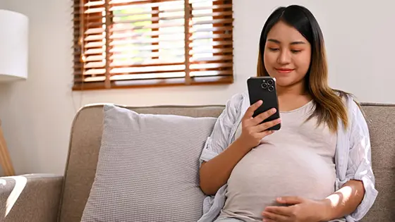 A young pregnant person sitting looking at their phone and with one of their hands resting on their tummy