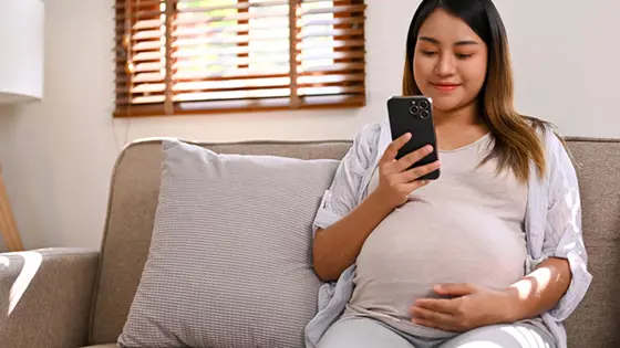 A young pregnant person sitting looking at their phone and with one of their hands resting on their tummy