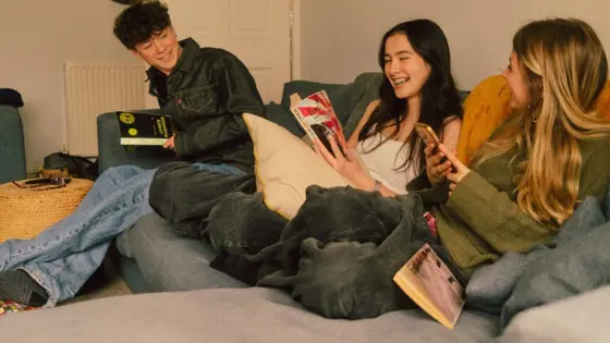 Photograph of three teenagers sat on a sofa, laughing, with one reading and one on their phone.