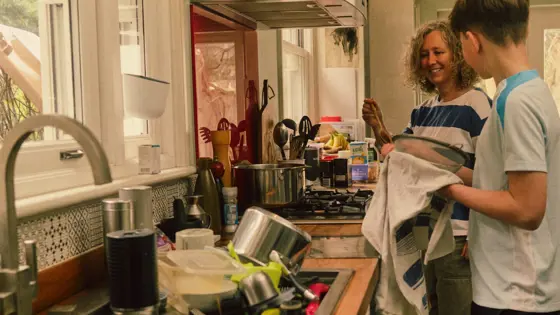 A carer smiling while washing dishes with a teenager.