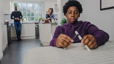 Photo of a teenager sitting at the table at home, with their parents out of focus in the background.