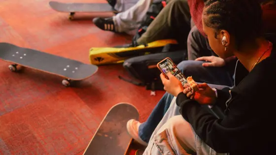 Photograph of teenagers sat inside with skateboards, using their mobile phones. 