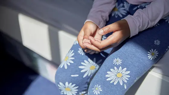A young person sitting on the edge of a bed, holding their hands on their lap. They seem unsure.