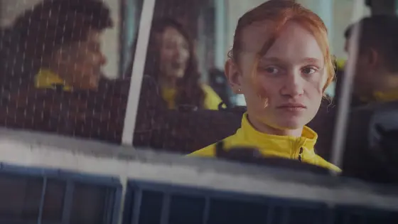 A young girl sat alone on a bus looks out of a window while other children talk in the background.