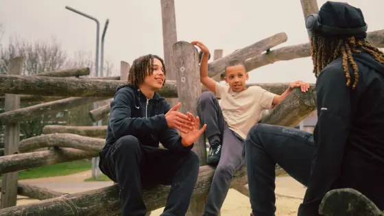 Photograph of three teenagers sat on a wooden structure outdoors.