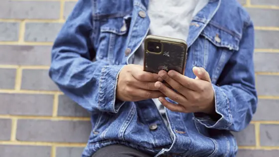 A young person leaning against a brick wall with their phone in their hands.